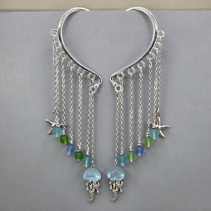 May include: A pair of silver-toned ear cuffs featuring multiple dangling chains. Each chain is adorned with small frosted blue and green beads, a silver starfish charm, and a larger translucent blue jellyfish-shaped pendant at the base.