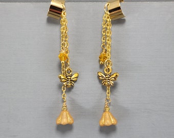 Könnte beinhalten: Ein Paar goldene Ohrclips mit baumelnden Anhängern. Jeder Ohrclip hat einen goldenen Bienenanhänger, einen kleinen goldenen Blumenanhänger und ein paar kleine gelbe Perlen.