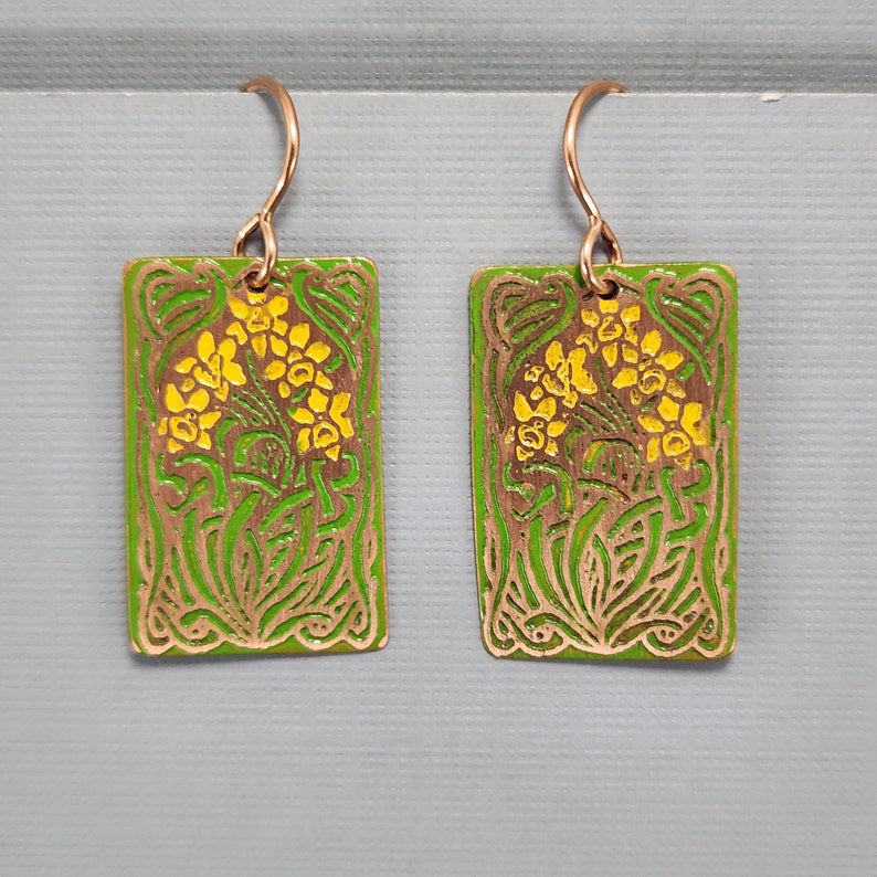 May include: A pair of green and gold earrings with a floral design. The earrings are rectangular and have a hook closure.