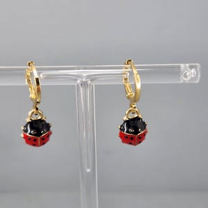 May include: A pair of gold hoop earrings with a dangling ladybug charm. The ladybug is red with black spots and a black head.