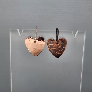 May include: A pair of heart-shaped earrings. One earring is a hammered copper with a shiny finish, and the other has a textured, dark brown finish. Both earrings have a simple hook design.