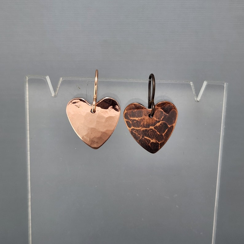 May include: A pair of heart-shaped earrings. One earring is a hammered copper with a shiny finish, and the other has a textured, dark brown finish. Both earrings have a simple hook design.