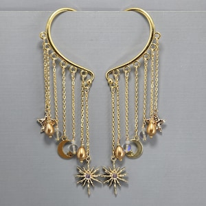 Könnte beinhalten: Goldfarbene Ohrklemmen mit herabhängenden Ketten, Sternen, Monden und tropfenförmigen Perlen. Die Ohrklemmen haben unten ein Sonnenstrahl-Design. Der Schmuck wird vor grauem Hintergrund präsentiert.