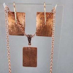May include: A copper necklace and earrings set with a square pendant and matching earrings. The necklace has a delicate chain and the pendant is attached to a small, ornate bail.