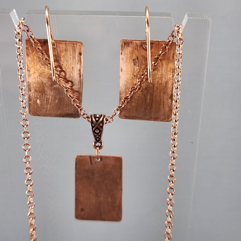 May include: A copper necklace and earrings set with a square pendant and matching earrings. The necklace has a delicate chain and the pendant is attached to a small, ornate bail.
