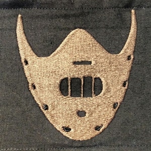 May include: Embroidered brown mask with a distinctive design, featuring a central opening with three vertical slots and a series of small holes along the edges. The mask has curved, horn-like extensions on each side, set against a dark background.