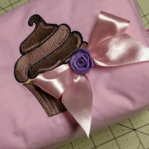 May include: A pink fabric bag with an embroidered cupcake design. The cupcake features brown frosting and a brown base. A pink satin bow and a small purple rose embellish the bag.