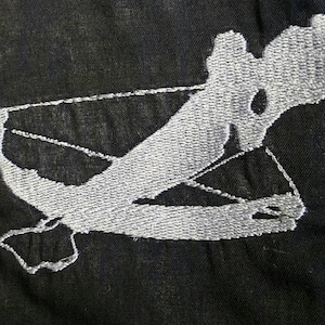 May include: A silver embroidered design of a bird in flight on a black fabric background.