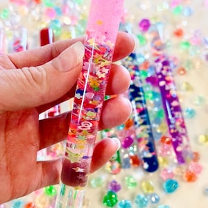 Puede incluir: Un tubo sensorial cil&iacute;ndrico transparente lleno de l&iacute;quido rosa, purpurina y formas coloridas tipo confeti. El tubo se sostiene en una mano, con otros tubos similares y cuentas de colores dispersas en el fondo.