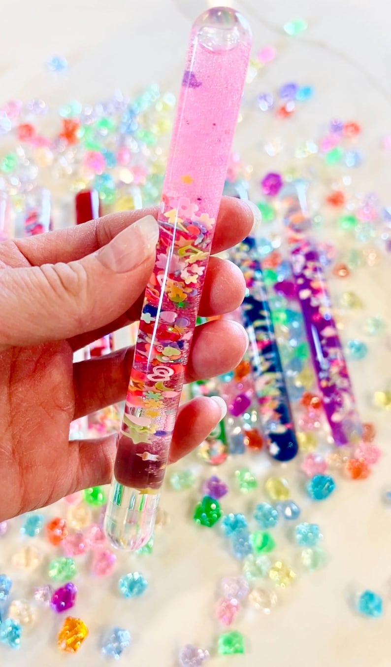 Puede incluir: Un tubo sensorial cil&iacute;ndrico transparente lleno de l&iacute;quido rosa, purpurina y formas coloridas tipo confeti. El tubo se sostiene en una mano, con otros tubos similares y cuentas de colores dispersas en el fondo.