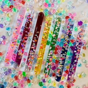 Puede incluir: Una variedad de tubos coloridos llenos de purpurina, cada uno con un dise&ntilde;o y esquema de color &uacute;nicos. Los tubos est&aacute;n dispuestos con una dispersi&oacute;n de peque&ntilde;as formas multicolores en forma de gema. Los tubos miden aproximadamente 15 cm de largo.