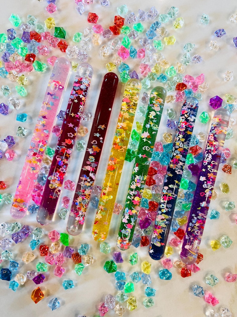 Puede incluir: Una variedad de tubos coloridos llenos de purpurina, cada uno con un dise&ntilde;o y esquema de color &uacute;nicos. Los tubos est&aacute;n dispuestos con una dispersi&oacute;n de peque&ntilde;as formas multicolores en forma de gema. Los tubos miden aproximadamente 15 cm de largo.