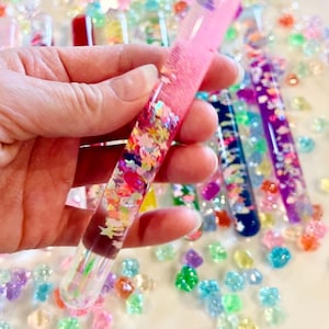 Puede incluir: Un tubo de vidrio transparente lleno de l&iacute;quido rosa y confeti colorido, sostenido en una mano. El tubo est&aacute; rodeado de cuentas dispersas, coloridas, con forma de gema. Otros tubos con l&iacute;quidos y confeti de diferentes colores son visibles en el fondo.