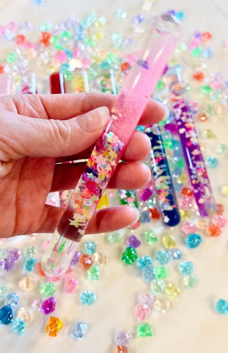 Puede incluir: Un tubo de vidrio transparente lleno de l&iacute;quido rosa y confeti colorido, sostenido en una mano. El tubo est&aacute; rodeado de cuentas dispersas, coloridas, con forma de gema. Otros tubos con l&iacute;quidos y confeti de diferentes colores son visibles en el fondo.