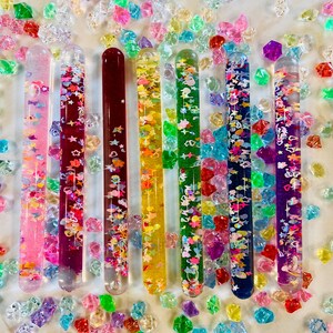 Puede incluir: Una colecci&oacute;n de tubos sensoriales coloridos llenos de purpurina y peque&ntilde;as formas, dispuestos sobre una superficie blanca salpicada de gemas acr&iacute;licas de colores. Los tubos son en tonos rosa, rojo, amarillo, verde, azul y morado.