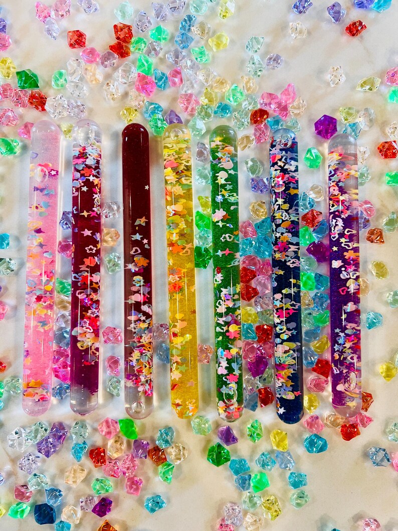 Puede incluir: Una colecci&oacute;n de tubos sensoriales coloridos llenos de purpurina y peque&ntilde;as formas, dispuestos sobre una superficie blanca salpicada de gemas acr&iacute;licas de colores. Los tubos son en tonos rosa, rojo, amarillo, verde, azul y morado.