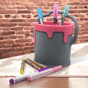 May include: A grey paint can shaped pencil holder with a pink rim and dripping paint design. The holder is filled with colourful markers and pens.