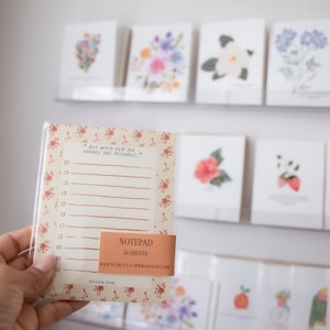 May include: A notepad with a floral design and the text "BUT WITH GOD ALL THINGS ARE POSSIBLE." The notepad has a list format and a peach-colored label that reads "NOTEPAD 56 SHEETS." Other floral cards are visible in the background.