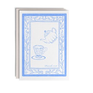 May include: A white greeting card featuring a blue ink illustration of a teapot and a teacup on a saucer. The design is enclosed in a blue floral border with a rectangular frame. The text "Proverbs 31:26" is printed in the bottom right corner.