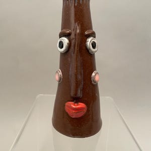 May include: A whimsical, brown ceramic vase with a face design. The vase features large, white-rimmed eyes, pink cheeks, and red lips. The vase is approximately 7 inches tall.