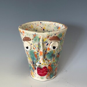 May include: A ceramic cup featuring a sculpted face with brown eyebrows, white eyes, and glossy red lips. The surface is decorated with a colorful speckled pattern in shades of orange, blue, and green over a cream-colored base, creating a textured look.
