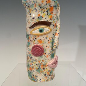 May include: A cylindrical ceramic vase with a white base featuring a multi-colored paint splatter pattern in orange, blue, and yellow. The vase has a sculpted face with a brown eyebrow, a single eye, a textured pink circular cheek, and pink lips.