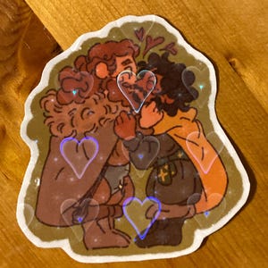 Op de afbeelding: Een sticker met een cartoonillustratie van twee mensen die elkaar omhelzen. De personages zijn getekend in een grillige stijl met bruine en oranje tinten. De achtergrond is groen en bruin met een patroon van harten.