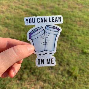May include: A sticker featuring two cartoon cups hugging, with purple liquid and smiling faces. The text "YOU CAN LEAN ON ME" is above and below the cups. The sticker is held against a green grassy background.