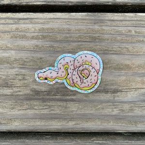 Op de afbeelding: Een holografische sticker met een slang ontworpen als donut. De slangvormige donut heeft roze glazuur, gele vulling en chocoladesnippers. De sticker ligt op een verweerd houten oppervlak.