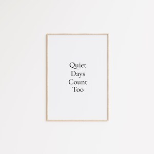 Può includere: Stampa incorniciata con le parole "Quiet Days Count Too" in un carattere nero ed elegante. Il testo è centrato su uno sfondo bianco, racchiuso in una cornice di legno marrone chiaro. L'opera d'arte è semplice e minimalista.