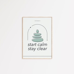 May include: A framed print with a minimalist design. The artwork features a stack of light green stones under a black arch, with the words "start calm stay clear" in a modern font. The frame is a light wood color.