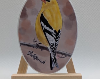 Puede incluir: Una impresión artística ovalada de un Jilguero. El ave es amarilla con alas negras y blancas, posada en una rama. La impresión tiene un fondo suave y se muestra en un pequeño caballete de madera. La palabra "Goldfinch" está escrita en la impresión.