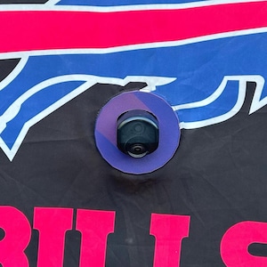 May include: A black and blue Buffalo Bills flag with a pink stripe and the word "BILLS" in pink letters. A security camera is mounted on the flag.