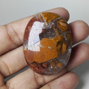 May include: Oval-shaped gemstone with a polished surface. The stone features a mix of colors, including shades of brown, orange, red, and clear quartz-like areas. The gemstone is held in a hand, showcasing its size and shape.