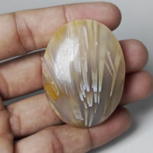 May include: An oval-shaped gemstone with a translucent, milky base. The stone features striking inclusions of golden-yellow and white, creating a pattern of radiating lines. The gemstone is held in a hand.