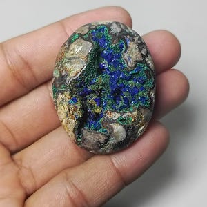 May include: Oval-shaped azurite stone with a mix of deep blue, green, and tan colors. The stone has a rough, textured surface with areas of crystalline formations. The stone is held in a hand.