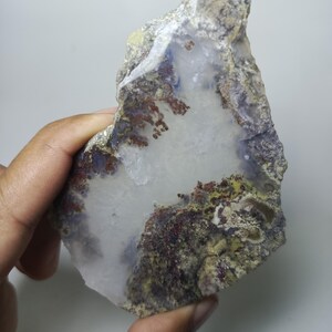 Puede incluir: Una losa de piedra natural en bruto con un centro blanco translúcido y un perímetro de colores mezclados. Los bordes muestran tonos de marrón, verde y morado, con inclusiones de color marrón rojizo. La piedra se sostiene en una mano.