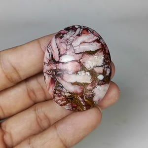 May include: Oval-shaped gemstone with a unique pattern of pink, white, and brown veins. The polished stone has a marbled appearance, with a mix of colors and textures. The gemstone is held in a hand.