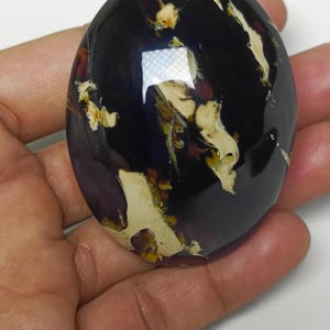 May include: Oval-shaped, polished amber stone with a deep purple hue, featuring creamy white and golden inclusions. The stone's surface reflects light, showcasing its smooth texture and natural patterns. A unique, organic gemstone.