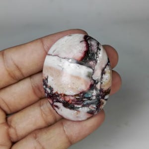 May include: An oval-shaped gemstone with a marbled pattern. The stone features a mix of colors, including white, pink, burgundy, and black. The polished surface of the stone has a smooth texture, and the colors are swirled together.