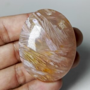 May include: An oval-shaped, polished gemstone with a translucent appearance. The stone features a blend of peach, yellow, and white hues, with delicate, feathery inclusions creating a unique pattern. The gemstone is held in a hand.