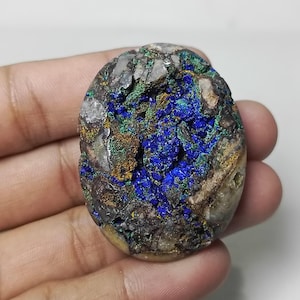 May include: An oval-shaped azurite and malachite specimen. The stone features a deep blue azurite center with green and brown accents. The outer edges are a mix of tan and gray, creating a natural, earthy appearance. The stone is held in a hand.