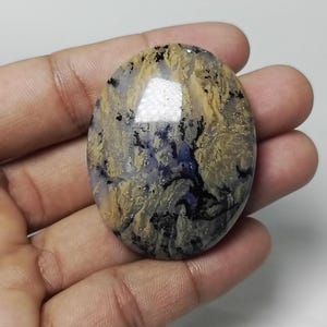 May include: Oval-shaped gemstone with a mix of colors. The stone features a blend of yellow, black, and purple hues, creating a unique pattern. The polished surface reflects light, highlighting the stone's natural beauty. The gemstone is held in a hand.