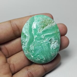 May include: A polished, oval-shaped gemstone with a vibrant green hue. The stone features a textured surface with areas of lighter green and white, creating a unique pattern. The gemstone is held in a hand.