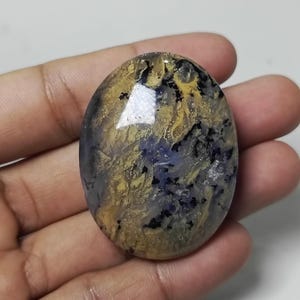 May include: An oval-shaped gemstone with a unique pattern of gold, blue, and black hues. The polished surface reflects light, highlighting the intricate details within the stone. The gemstone is held in a hand, showcasing its size and shape.
