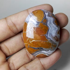 May include: An oval-shaped gemstone with a polished surface. The stone features a mix of colors, including shades of brown, orange, white, and gray. The pattern has a marbled appearance with visible cracks. The gemstone is held in a hand.