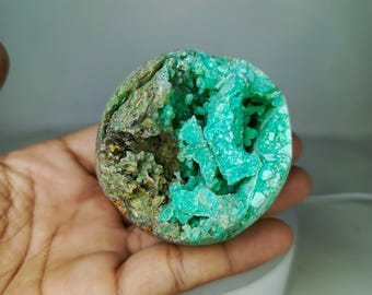 Puede incluir: Una geoda con un vibrante interior turquesa y un exterior áspero y terroso. La geoda se sostiene en una mano, mostrando su forma natural y orgánica. El interior presenta formaciones cristalinas, creando un contraste llamativo.