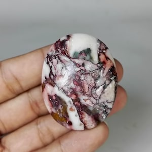 May include: Oval-shaped gemstone with a marbled pattern of white, burgundy, and gray. The polished stone has a smooth surface and a natural, organic appearance. The gemstone is held in a hand.