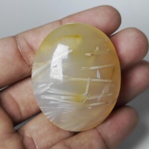 May include: Oval-shaped gemstone with a translucent, yellow-orange hue. The stone features white, feathery inclusions that create a unique pattern. The polished surface reflects light, highlighting the natural variations within the stone.