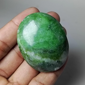 May include: A smooth, oval-shaped green stone with white and darker green marbling. The polished surface reflects light, highlighting the natural patterns. The stone is held in a hand, showcasing its size and texture.